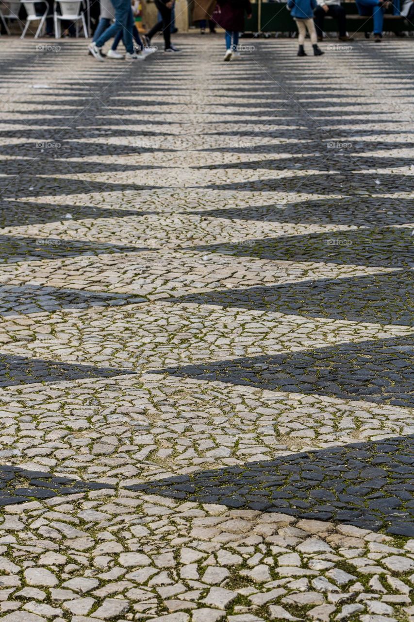 Black and white cobblestones in a chequered pattern lead to people in the distance, walking by or sitting outside cafes