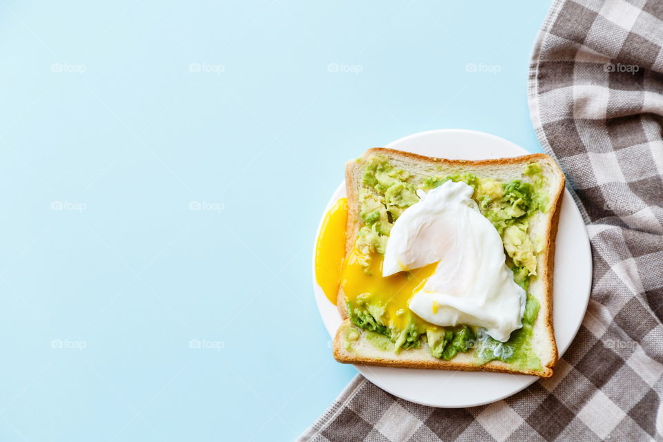 Sandwich with fresh green avocado, poached egg on white plate lying on blue background 