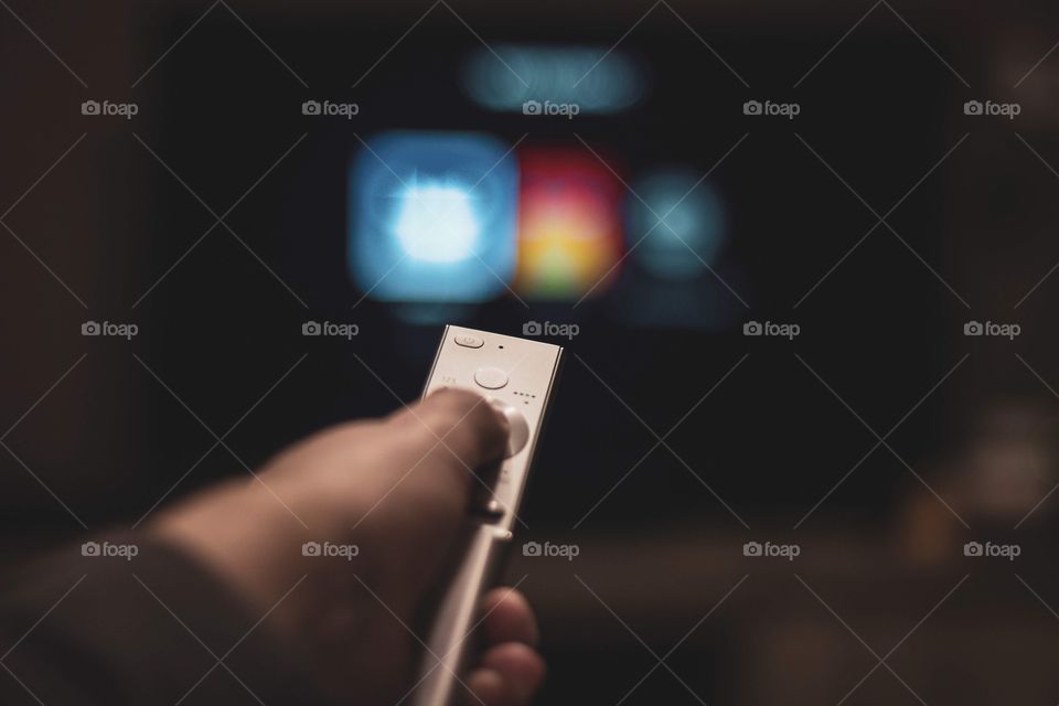 A portrait of a the hand of a person holding a television remote control. in the blurred background you can see the user selection screen of Netflix.