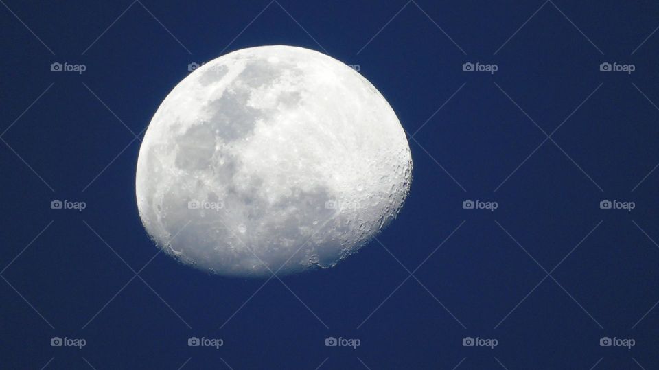 Unusual shot of bright white moon taken during the day against a deep blue sky with moon craters and seas clearly visible 