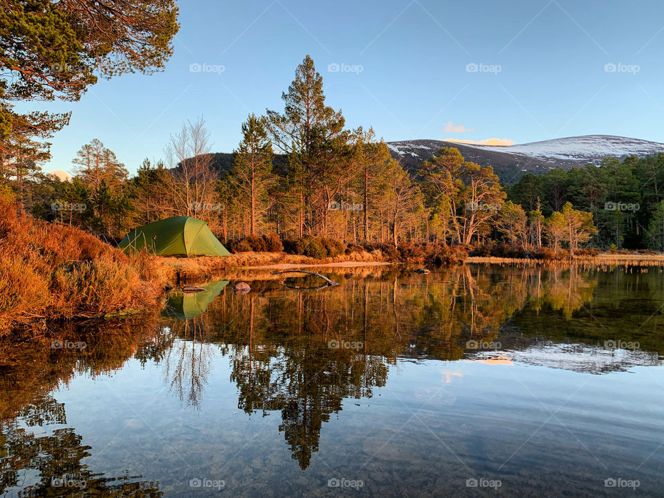 Wild camping in the Cairngorms, Scotland, a popular hiking destination in the Scottish Highlands. A still lake reflects the scene with a tent, snowy mountains and pine forest.