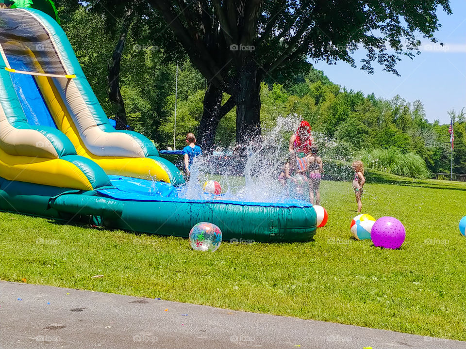 child splashing into inflatable slide pool while other children admire and chat with Ariel the mermaid at a birthday party.