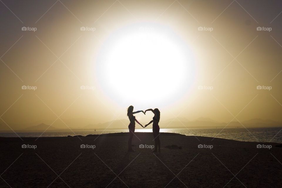 Two girls forming a heart with the sun in the background.