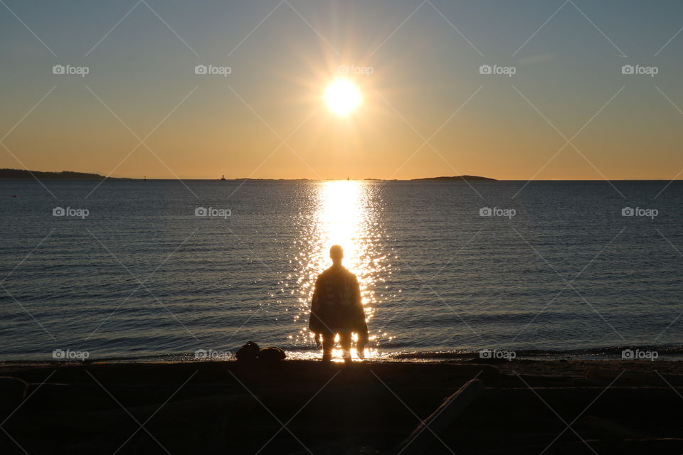 Early morning on the beach with sun rising up slowly above the ocean and reflecting in it as thousands little bright stars .. A young  man  wearing a towel stands still adoring this beautiful morning full with tranquility and simple joy ...