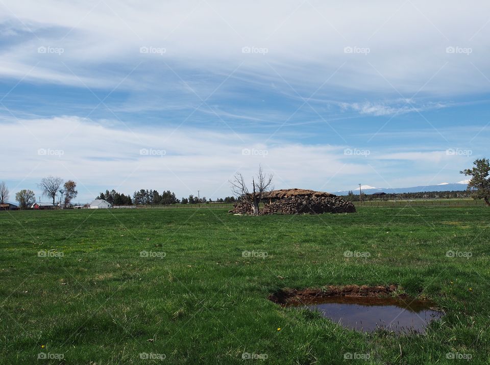 An old homesteading cold storage building built from rocks in the field with mountains in the background and water hole in the foreground on a sunny spring morning. 