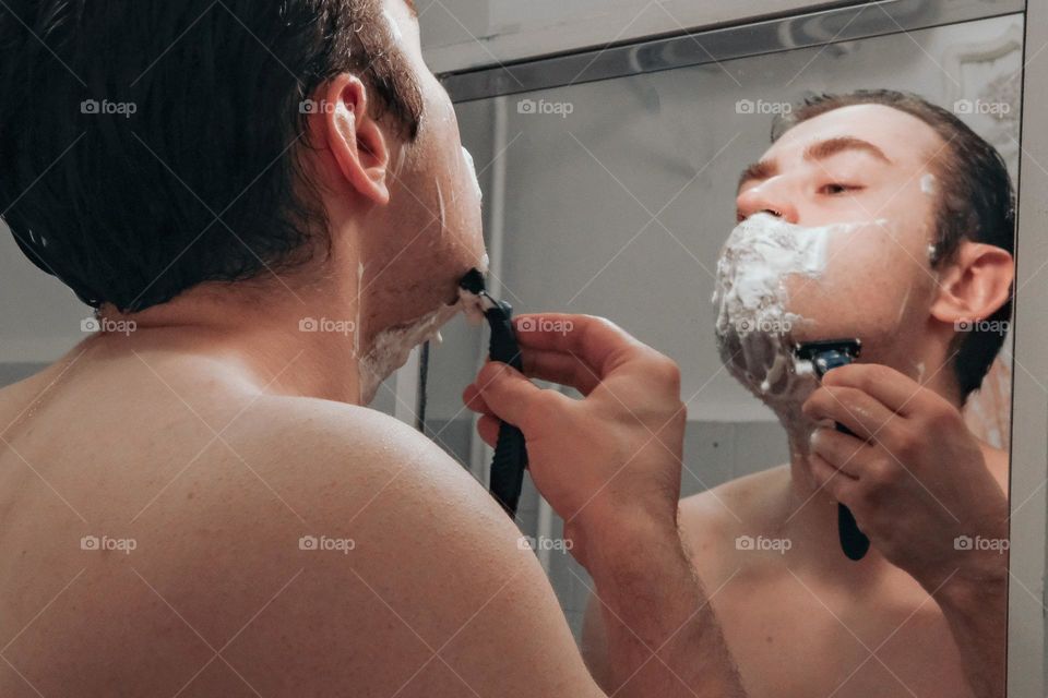 A man shaves his beard while looking in the mirror