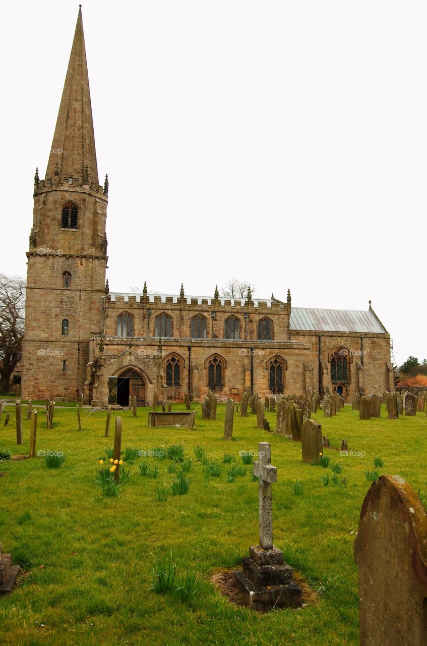 The churchyard in Masham, UK.  Old headstones and daffodils sit before a historic church.  Grey sky.