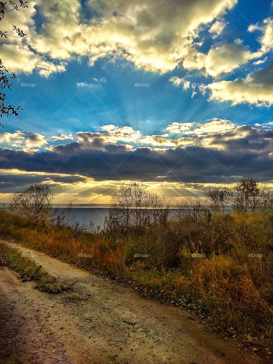 Very beautiful view of the Volga River (Russia) and the horizon on the opposite bank, footpath, bushes and trees covered with golden foliage in the foreground, under the blue sky covered with clouds and rays of the setting sun