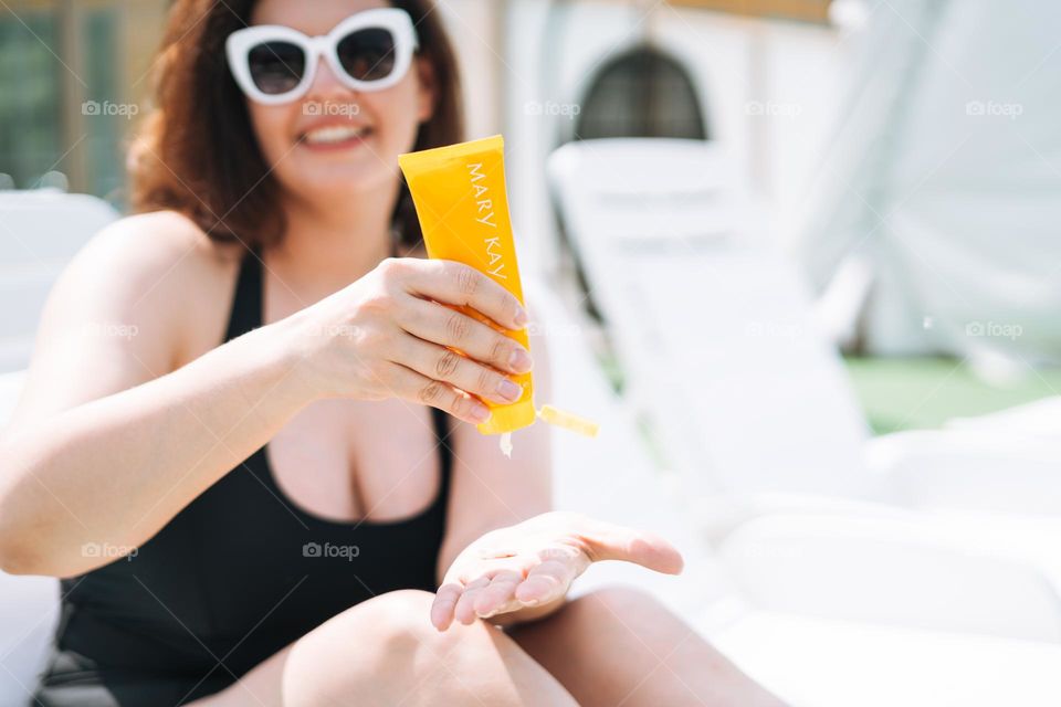 Stylish happy young woman plus size body positive in black swimsuit and sunglasses with sunscreen cream Mary Kay in hands on beach lounger, summer vacations
