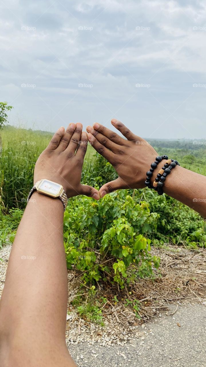 Hands form a triangle by touching thumbs and index fingers, directed towards lush green vegetation.