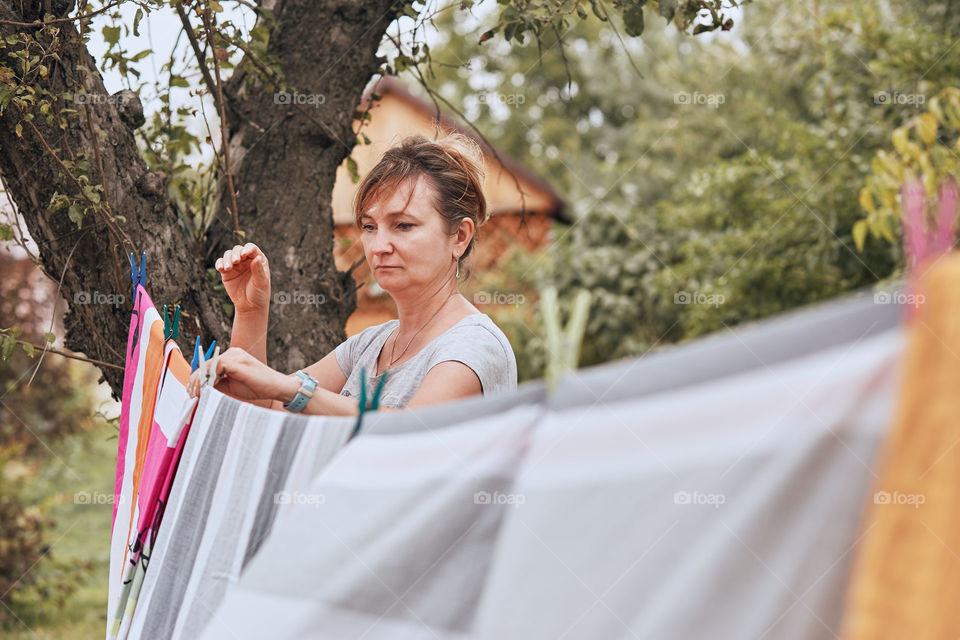 Mature woman hanging a freshly laundered bed linen on clothesline stretched between two trees in a orchard. Candid people, real moments, authentic situations