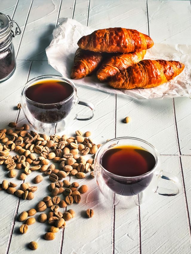 Two cups of hot black coffee, crispy croissants and coffee beans scattered at the side
