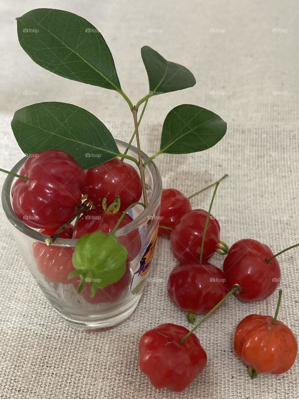 Copo cheio de frutinhas vermelhas de pitanga. Uma frutinha verde e galho com folhas. Composição feita em cima da mesa.