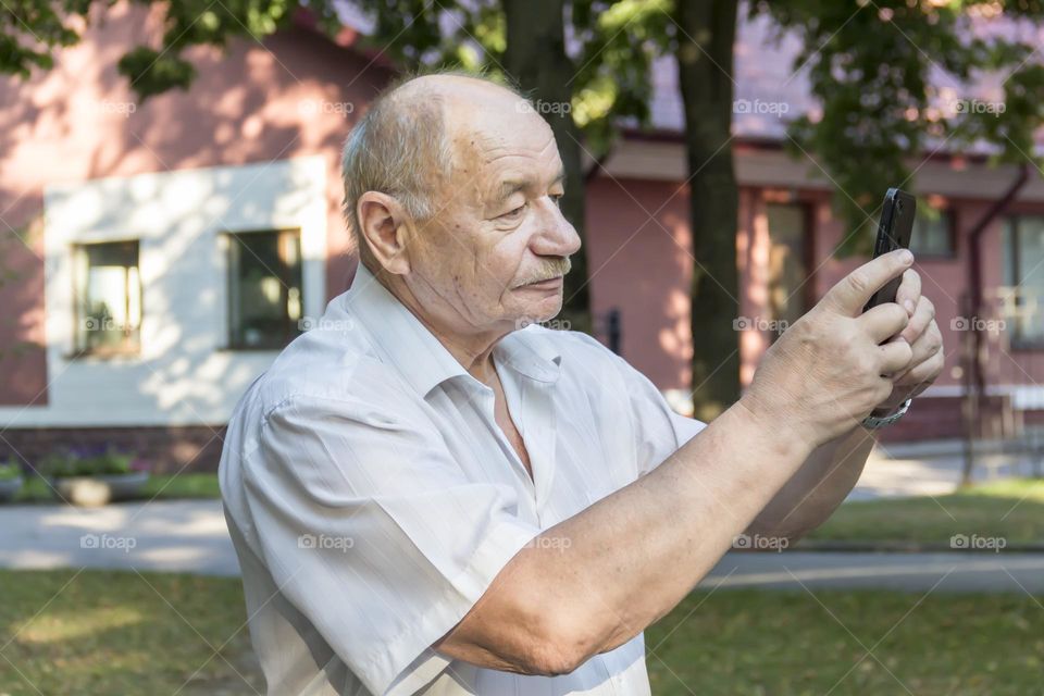 An elderly man walks alone in the park in the summer. A modern pensioner, businessman in a white shirt and trousers takes pictures with a camera in a mobile phone.