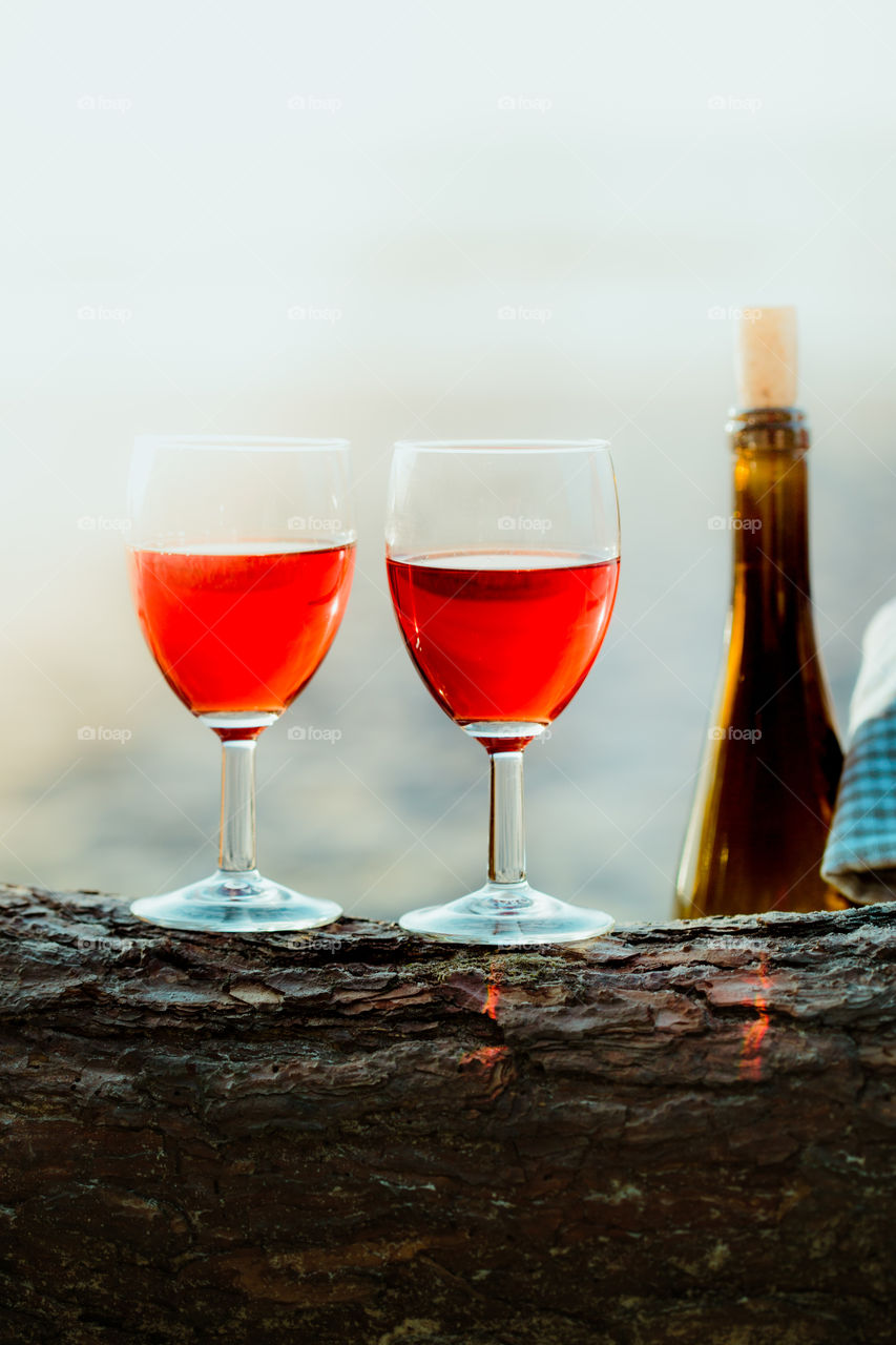 Two wine glasses with red wine standing on tree trunk, on beach, beside wicker basket with bottle of wine