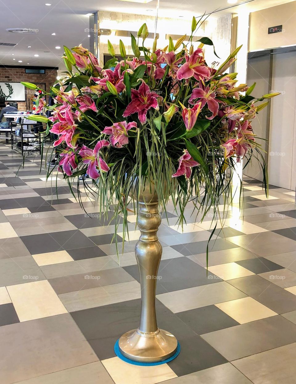 Lush bunch of beautiful purple pink lilies with green leaves in the narrow tall bronze color jar vase in the middle of the building hall