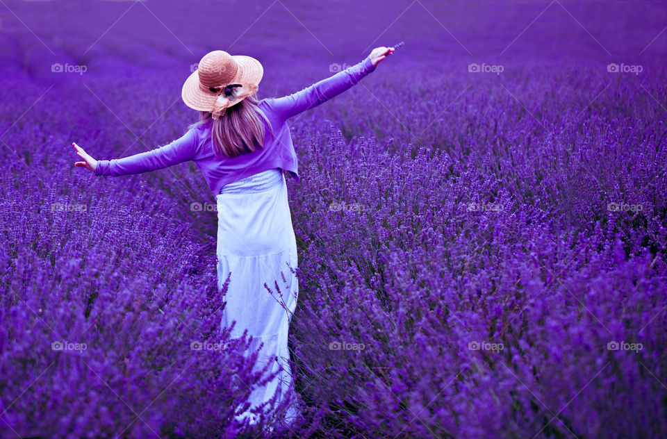 Back view of young blond woman in lavender field. Happy and carefree female in a white dress and straw hat enjoying nature. Outdoors portrait.