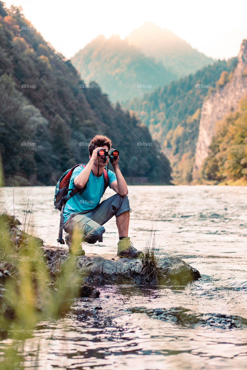 Young tourist with backpack looks through a binoculars on mountains peaks, stands on a rock over a river. Boy spends a vacation in mountains, wandering with backpack, he is wearing sports summer clothes