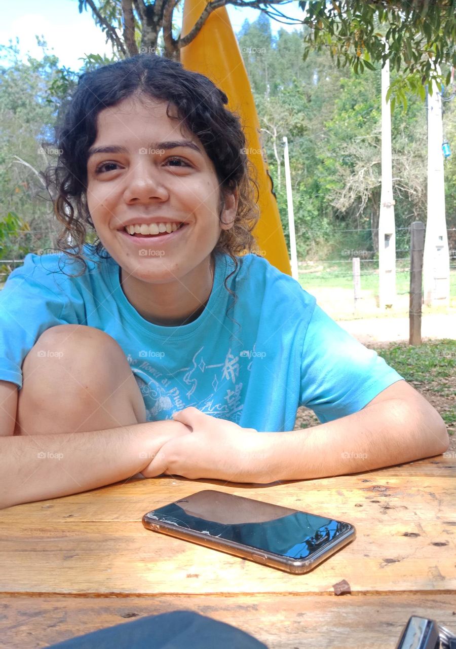 Young girl, sitting and smiling with arms over a table. Curly hair,  blue t-shirt, natural background with trees,  a phone on the table