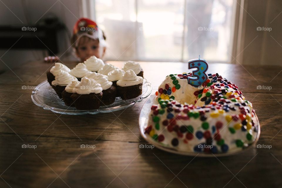 Three year old child looking longingly at a birthday cake. Staring over table at birthday cake and cupcakes