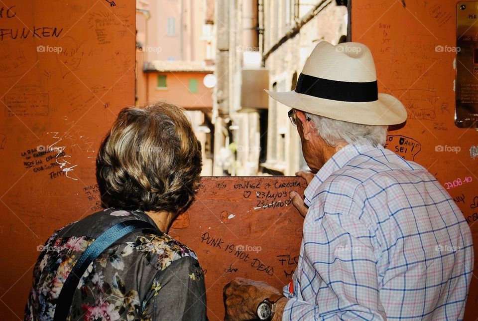 elderly couple observe a residential neighborhood through a window carved into a wall in Bologna, Italy