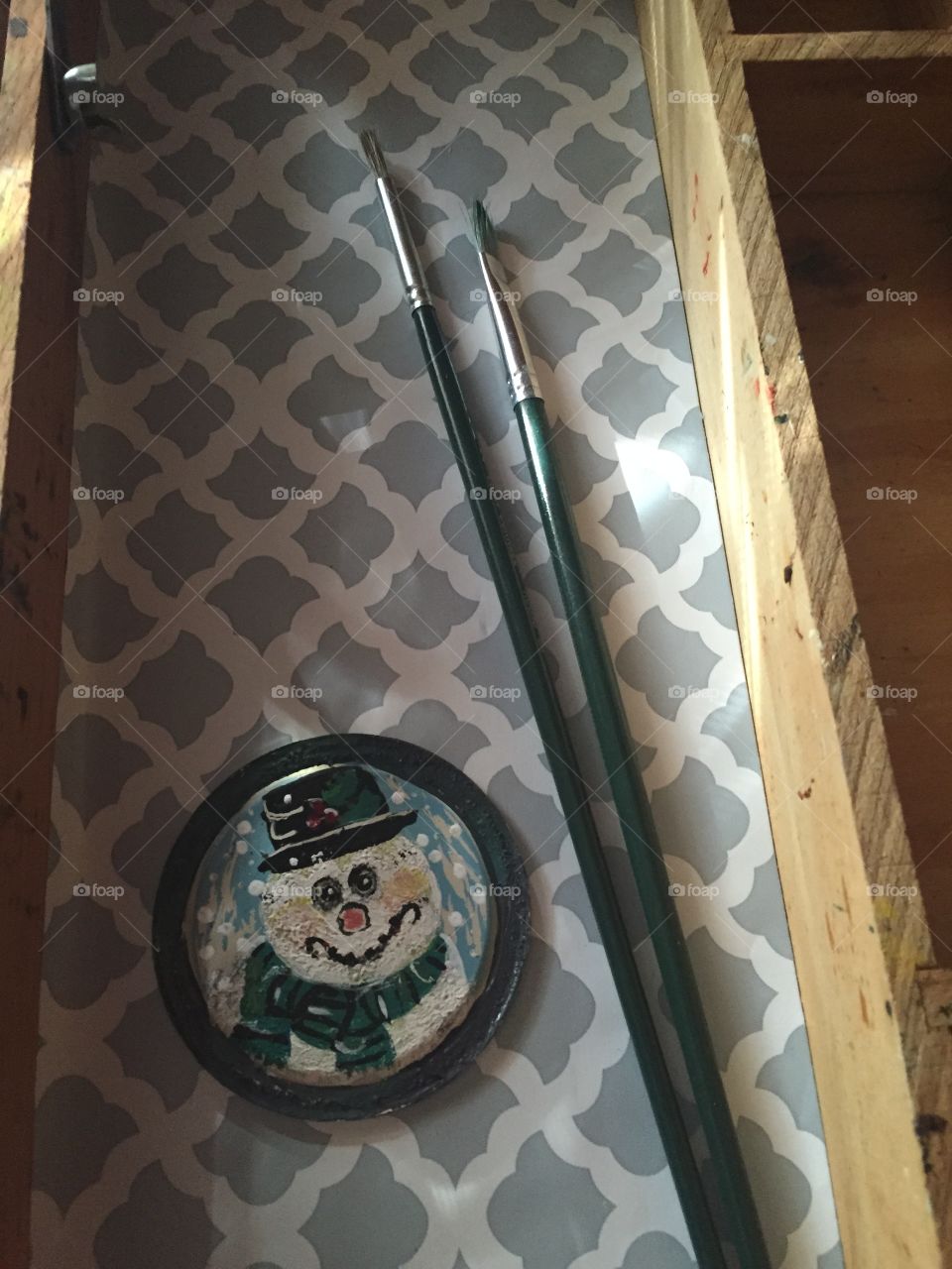 Snowman face with black hat and green scarf  ornament painted on a small circular disc with my paintbrushes in sight and sitting in my wooden paintbox. 