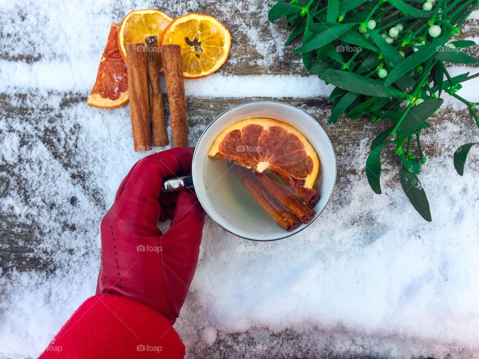 Woman’s hand wearing red glove holding cup of orange tea with mistletoe placed beside