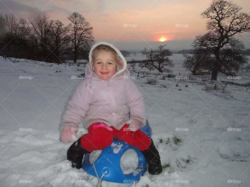 beautiful child sat on a sledge surrounded by British snow and setting sun .