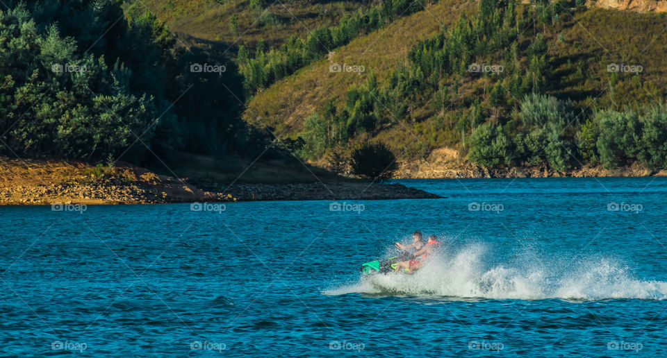 2 people on a jet sky, make a sharp turn spraying white water around them, on a calm blue river with green hills behind them