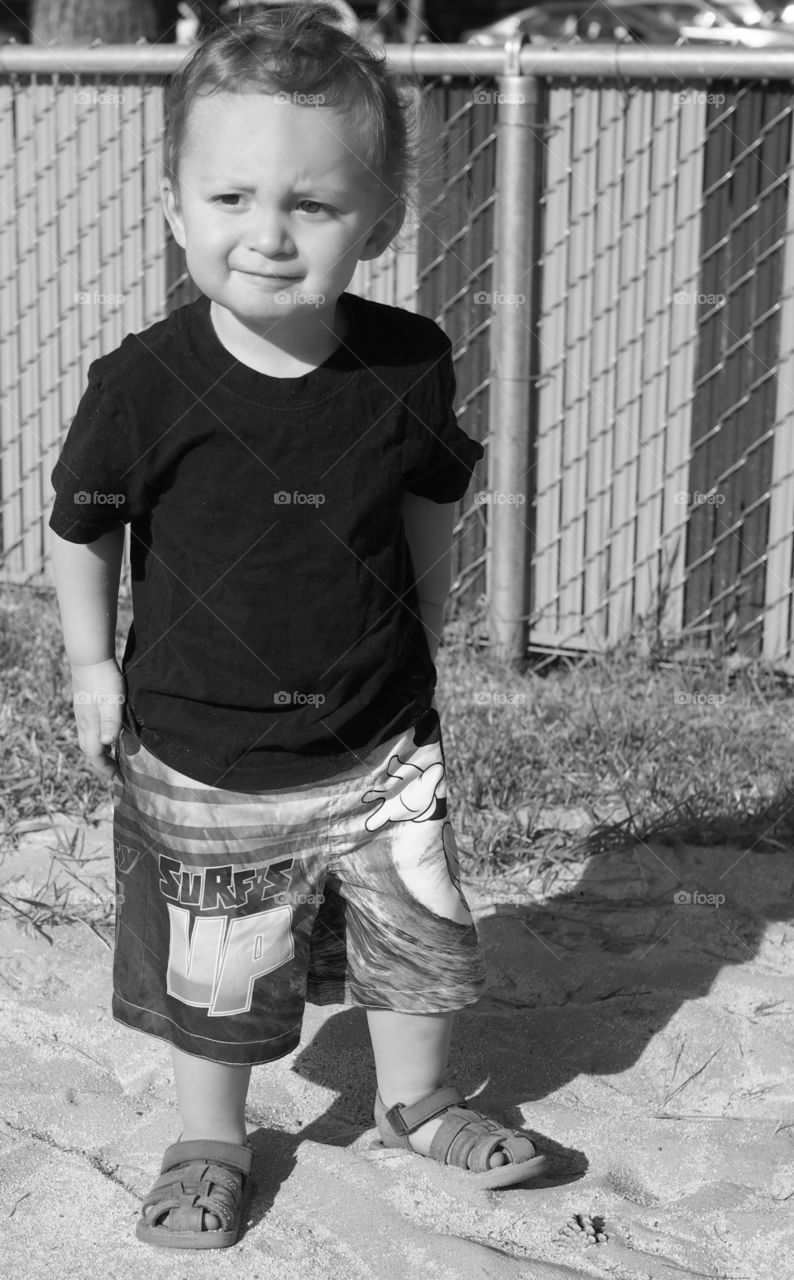 A small boy in shorts, t-shirt, and sandals stands in a sand pit with focused attention. 