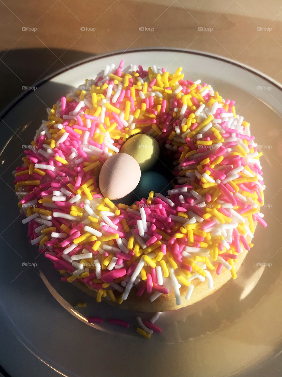 An Easter iced doughnut with pink,yellow and white sprinkles and pastel-coloured candy-coated chocolate eggs in the hole. Yummy! 🐣