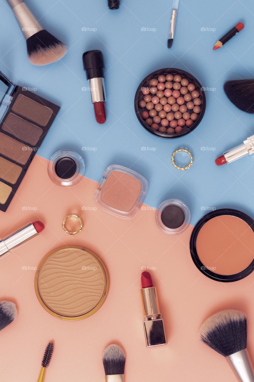 Set of women's cosmetics of eyeshadow, face powder, red lipstick, brushes, brushes and ring lie scattered on a pink and blue background, flat lay. Concept cosmetics female, beauty salon, makeup items.