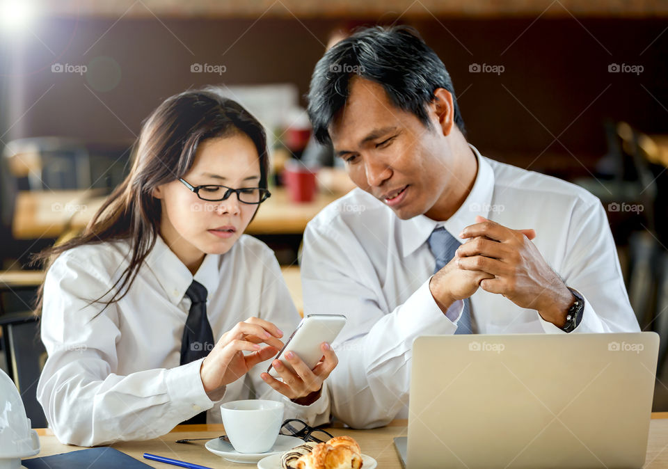 Two asian persons discuss business on mobile at meeting with laptop and coffee on table