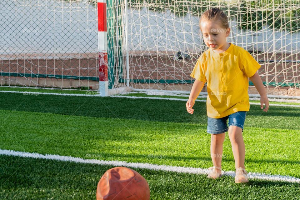 Cute little girl preparing kick soccer ball on green sports field at sunset time. Kid playing football with her family