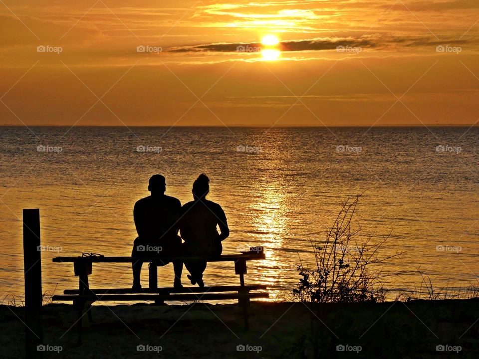 A romantic couple sitting on a picnic table watch a magnificent sunset descending over the shimmering bay.