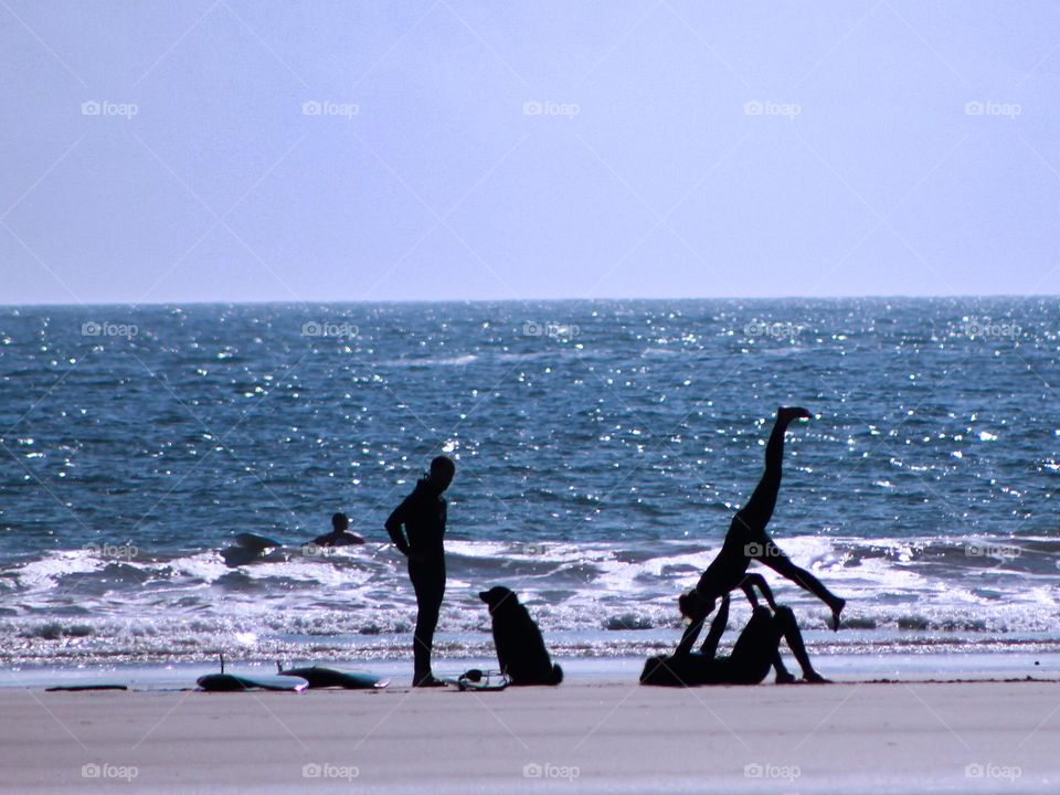 Silhouettes of people on the beach & in the beautiful blue ocean. People are body surfing with their boogie boards & there are more boards on the beach. One person is standing with their dog & one person is doing handstands on another’s knees. Fun!