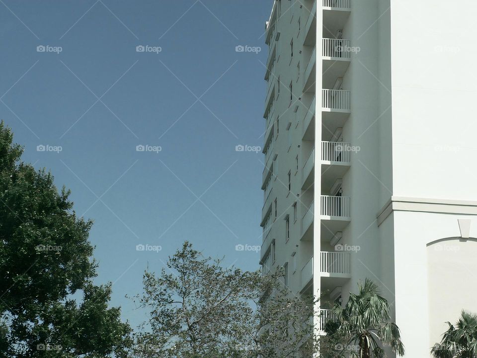 Symmetrical apartment’s units creating a geometrical effect with light and shadows by a clear blue sky and green leaves trees.