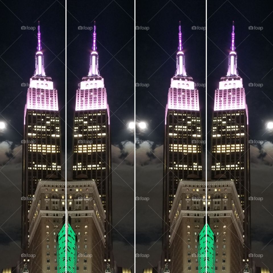 Abstract of the Empire State Building.  Photo taken at night in New York City. The moon is round & bright. The building is tall & lit at the top with purple and pink lights.The lights in the middle of the building are off white & green at the bottom.