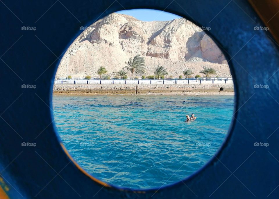 View through a boat's porthole of turquoise waters with desert mountains and palm trees along the shoreline