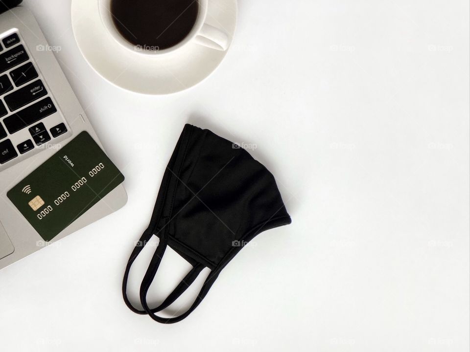 Laptop keyboard, credit card, black face mask, a cup of coffee. Top view, copy space.