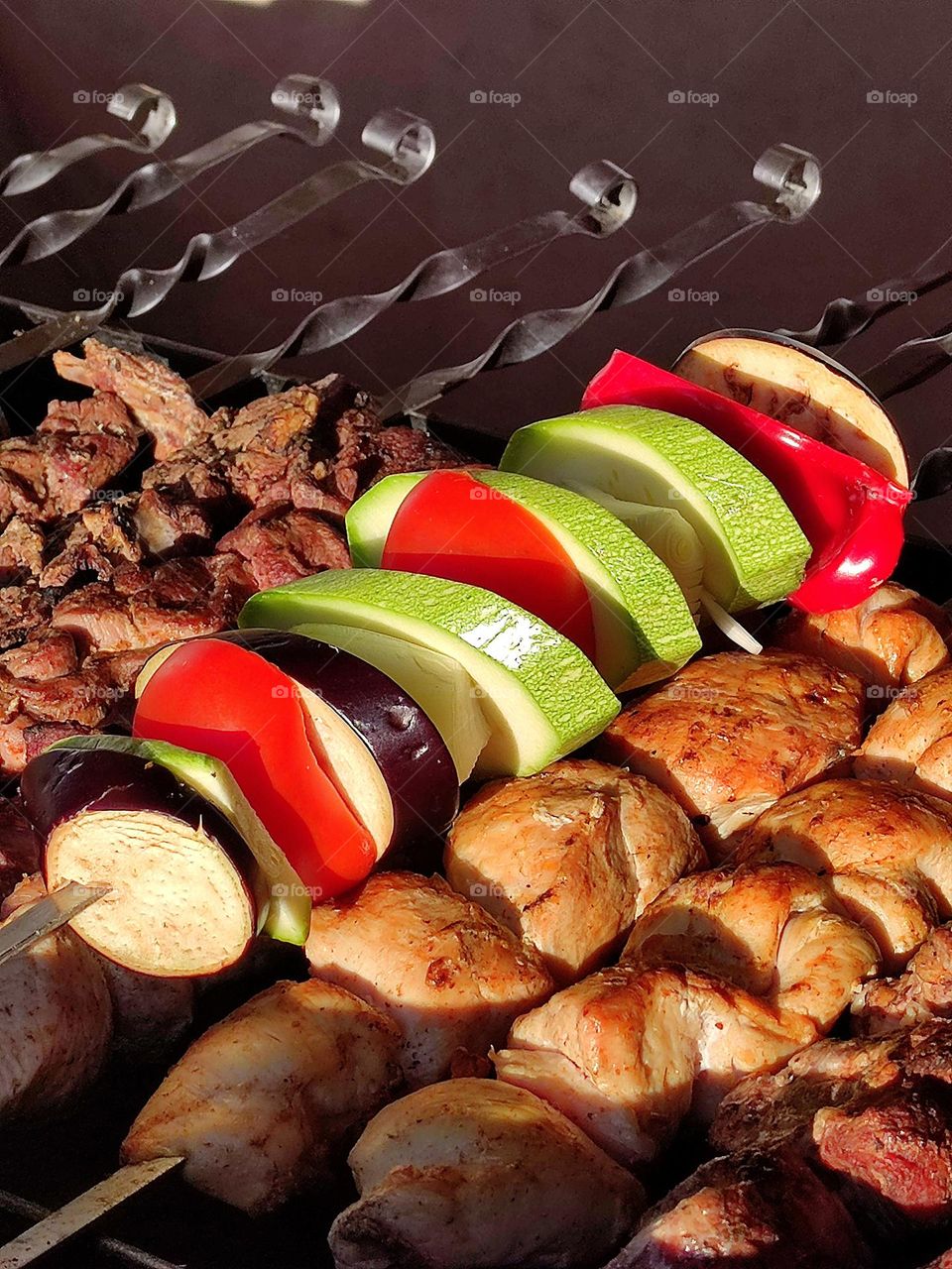 Grill. Grilled meat on metal skewers. Rings of purple eggplant, red bell pepper, green zucchini and onion are strung on one skewer
