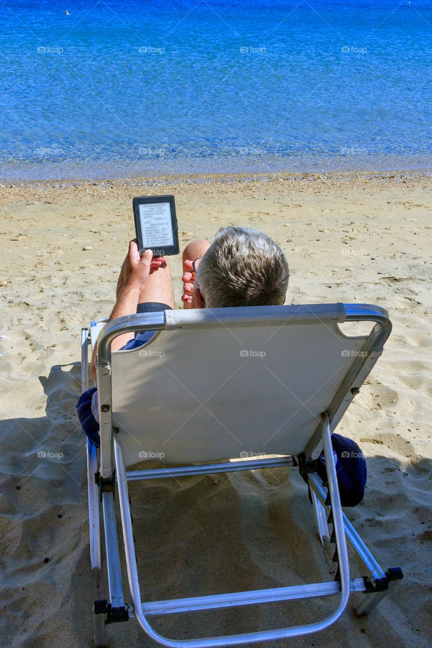 Back view of man reading electronic book on the beach