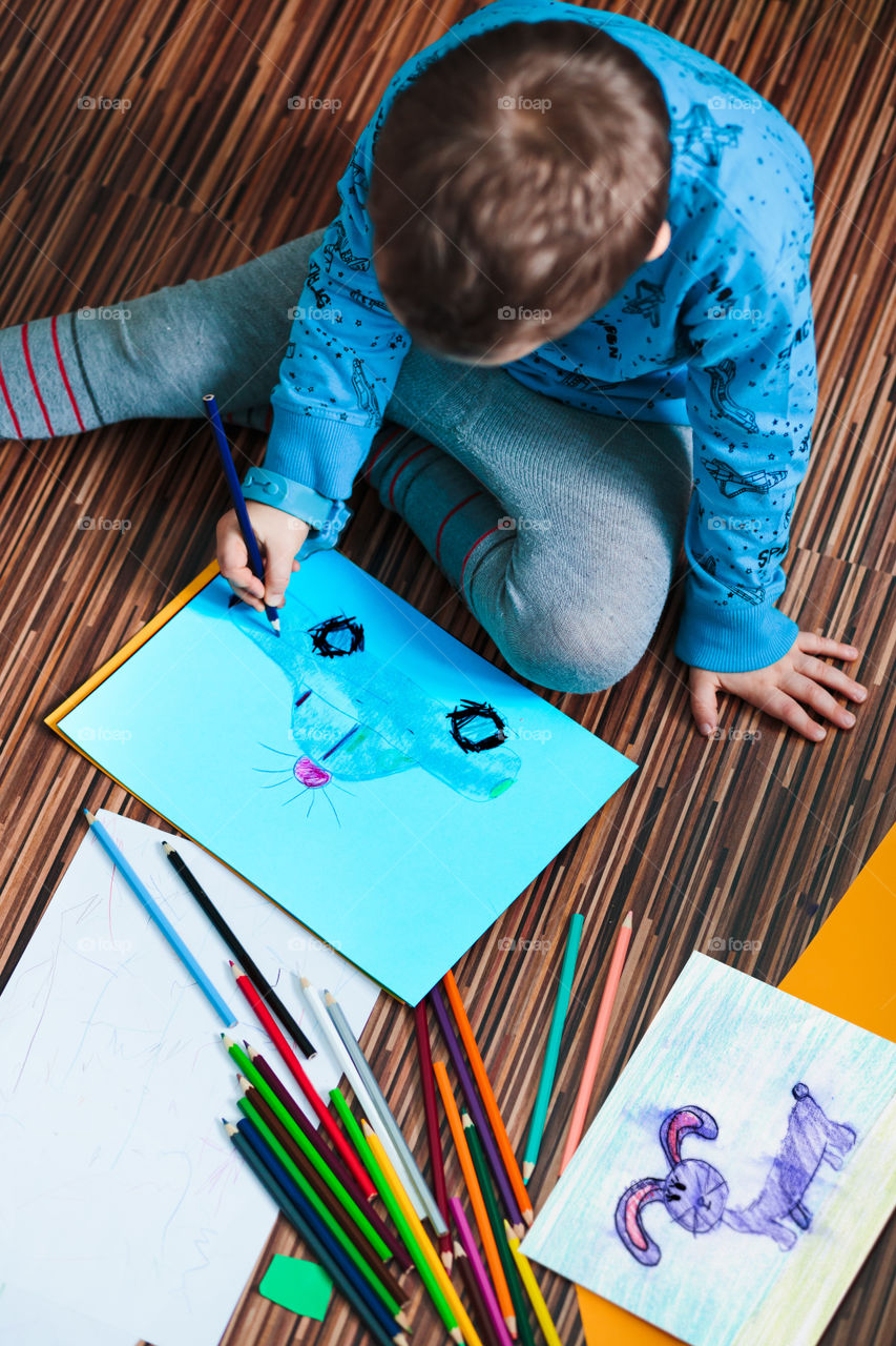 Little boy drawing a colorful picture of a car using pencil crayons sitting on floor. Shot from above