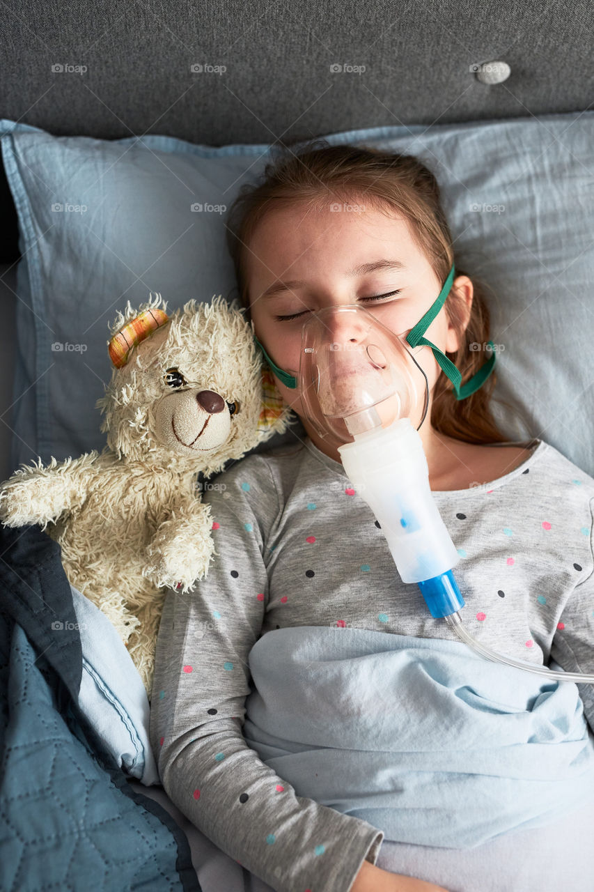 Sick little girl having medical inhalation treatment with nebuliser. Child with breathing mask on her face lying in bed