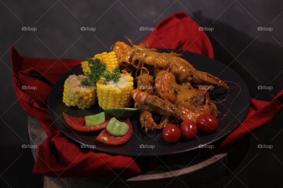 Asian food balado fried prawns with corn chunks, lemon slices and tomatoes served on black plate with red cloth bottom and wooden coaster. Asian style street seafood.