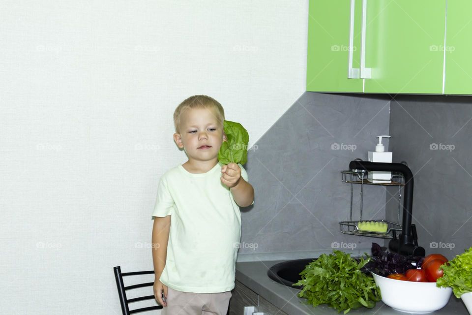 A small child washes fresh, green, vegetarian vegetables under the tap in a black sink to prepare salads and other dishes in a homemade gray and green kitchen.
