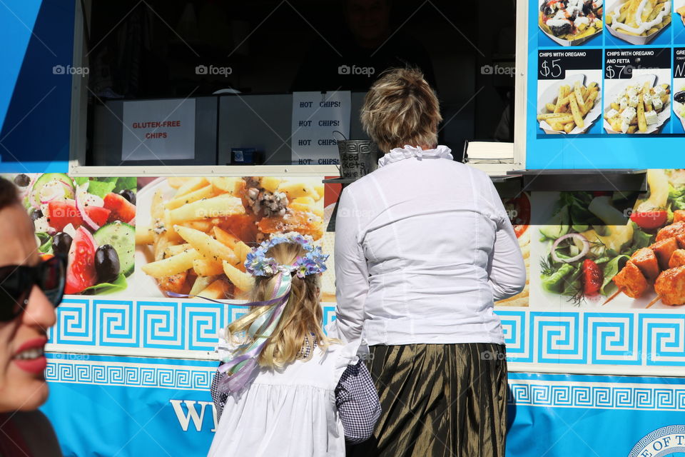 May Day may pole Cornish costumes. Little girl in checked dress and floral crown at outdoor festival food concession, back view