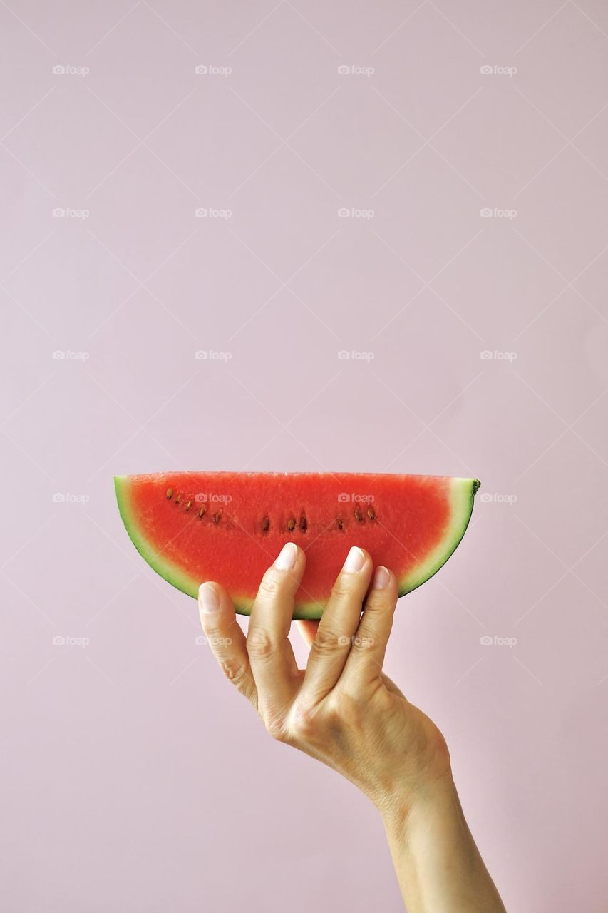 Cropped hand holding a slice of watermelon against pink background. 