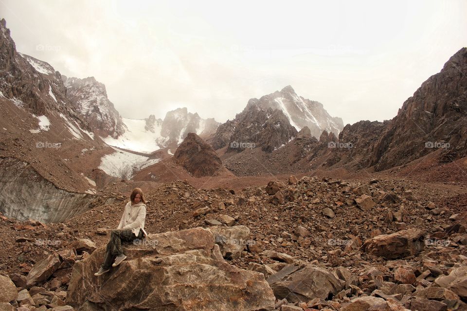 Girl in a boho knitted sweater sits on a large boulder on a glacier in the mountains