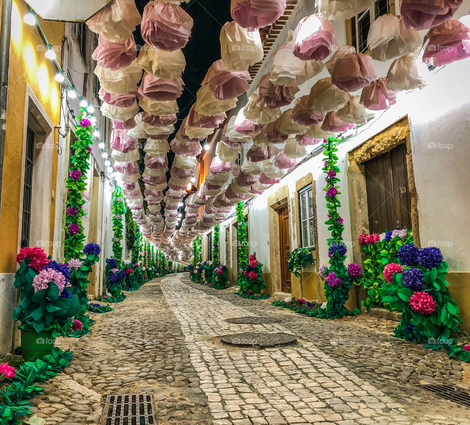 A nighttime, low view down a cobbled street decorated with colourful paper flowers and bright lights